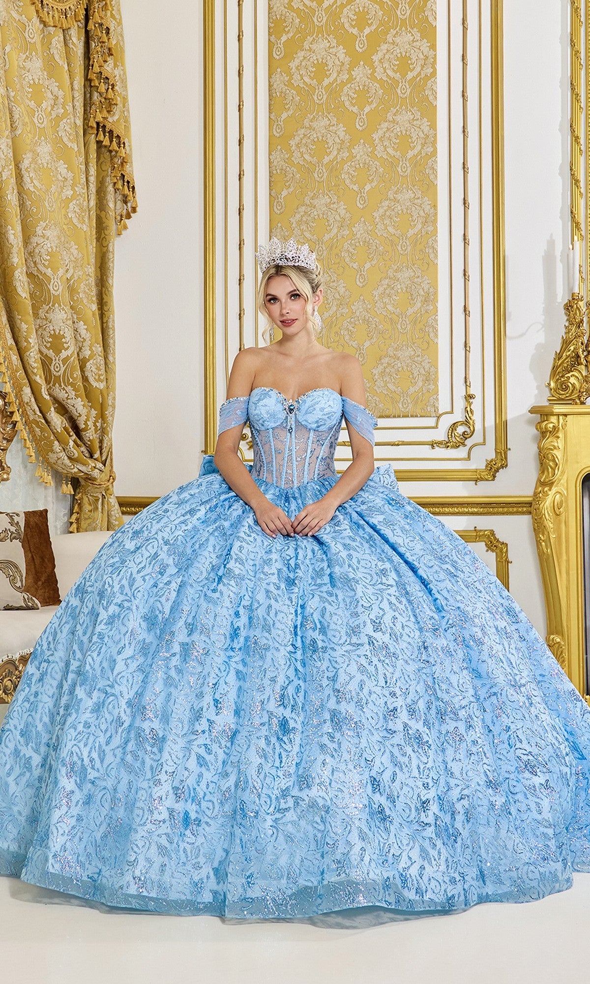A woman wearing the Quinceanera Dress 5095 By Dancing Queen in Bahama Blue and a tiara sits in an opulent room with gold and white decor.
